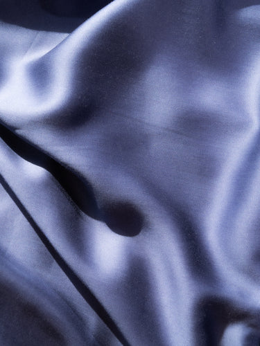 A close-up of draped satin fabric with soft folds and a subtle sheen, shown in a cool blue-violet tone against a matching light background. #color_denim