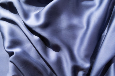 A close-up of draped satin fabric with soft folds and a subtle sheen, shown in a cool blue-violet tone against a matching light background. #color_denim