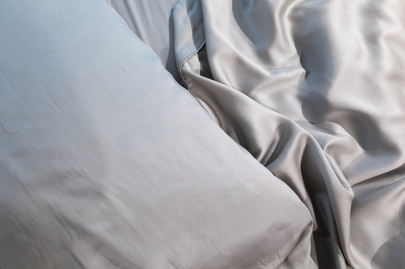 Close-up of light gray satin bedding, showing a smooth pillowcase and draped fabric with soft folds and a subtle sheen.  #color_silver